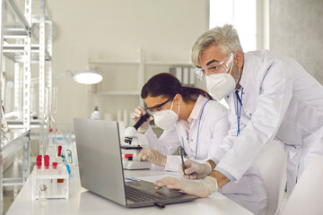 Microbiologist biotechnology researcher group in facial mask and protective gloves conducting experiment. Male supervisor making notes on laptop and female assistant analyzing sample using microscope