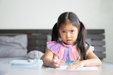 asian child back to school or kid girl made curious face to write or draw and read on book with doing homework to new idea think or people learn from home in bedroom and study online with book stack