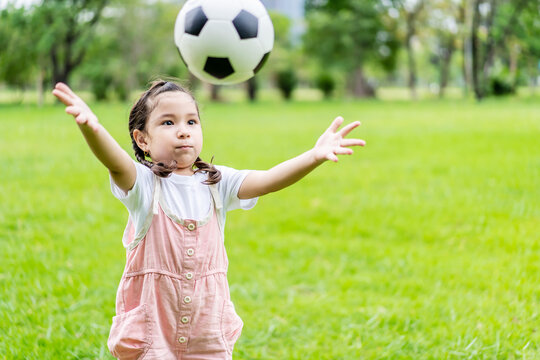 Smiling little girl standing throw the soccer ball at green football field in summer day. Portrait of little girl athlete playing with a ball at stadium. Active childhood concept. Copy space