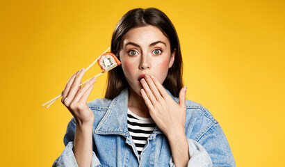 Girl holds sushi with chopsticks, looks surprised with special delivery offer, bonuses for takeaway orders in japanese restaurant. Woman amazed by delicious taste of sushi rolls, yellow background