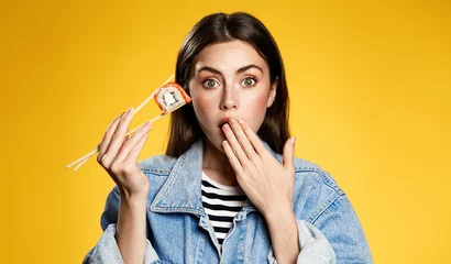 Tuinposter Girl holds sushi with chopsticks, looks surprised with special delivery offer, bonuses for takeaway orders in japanese restaurant. Woman amazed by delicious taste of sushi rolls, yellow background © Liubov Levytska