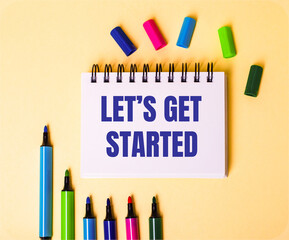 The words LET IS GET STARTED written in a white notebook on a beige background near multi-colored markers