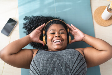Happy curvy African woman having fun listening music with headphones while doing pilates at home