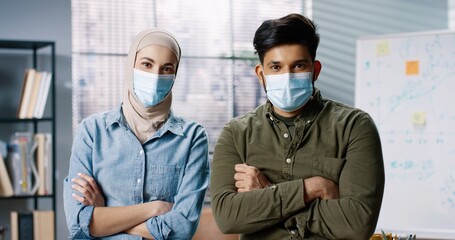 Fototapeta na wymiar Portrait of Arab young beautiful woman and Hindu man in medical masks in office looking at camera at workplace. Female and male employees in cabinet in quarantine, occupation concept