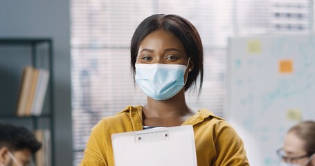 Fototapeta na wymiar Close up portrait of young cheerful African American female employee in medical mask standing at office, looking at camera and smiling. Joyful woman manager in good mood, coronavirus concept