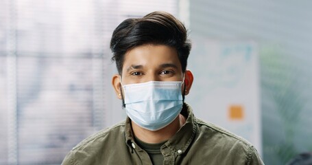 Close up portrait of young cheerful Hindu male employee in medical mask standing at office, looking at camera and smiling. Joyful handsome man manager in good mood, coronavirus concept