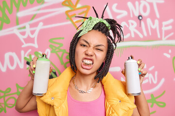 Youth and street lifestyle concept. Cheeky dark skinned woman with braids clenches golden teeth wears yellow vest metal chain around neck holds aerosol bottles draws graffiti wall in public place - obrazy, fototapety, plakaty