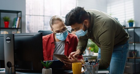 Fototapeta premium Portrait of Hindu male worker in medical mask come to Caucasian young man colleague and showing something on tablet device discussing something at workplace. office concept, company job