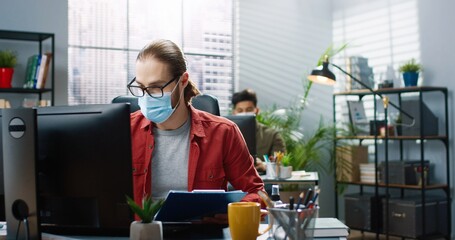 Fototapeta na wymiar Portrait of Caucasian handsome male worker in medical mask sitting at desk typing on computer. Woman colleague showing something to boss discussing business issues at workplace. office concept