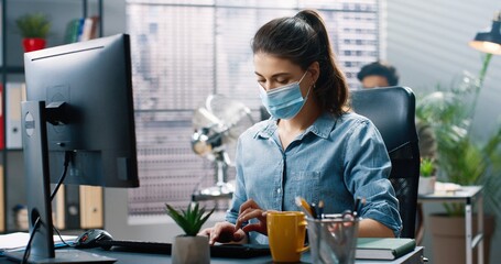 Close up portrait of beautiful young concentrated businesswoman in medical mask sitting at desk and...