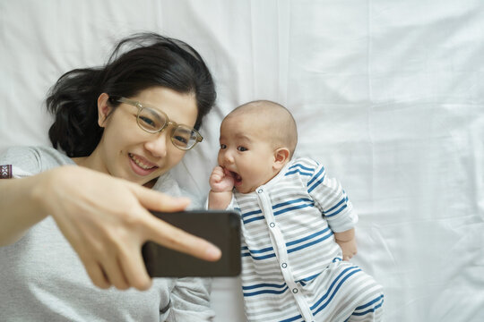 Asian Young mother and newborn baby son lying on bed having fun making video call and taking selfie photo on smartphone or mobile phone at home together