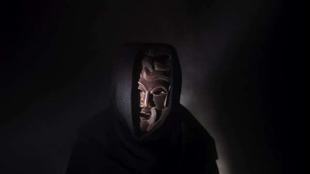 Creepy, masked cultist standing in a dark room. Scary horror scene, mysterious man in a tribal mask isolated on a black background. Eerie mask, devil concept.