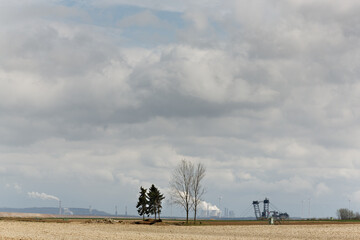 Ein Acker, 2 Nadelbäume. zwei kahle Laubbäume, ein Bagger im Braunkohletagebau Garzweiler sowie ein qaulmendes Kraftwerk , Windräder, eine Fabrik und Schornsteine vor Wolken und blauem Himmel