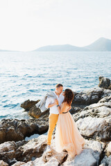 Smiling groom hugs the bride in a beautiful beige dress standing with her back on a rock above the sea