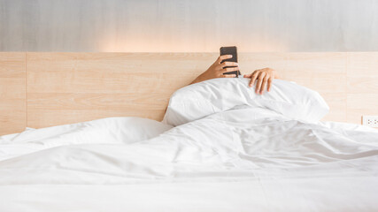 Man waking up and use smartphone or mobile phone on the bed in white blanket at morning time