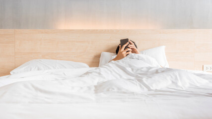 Man waking up and use smartphone or mobile phone on the bed in white blanket at morning time