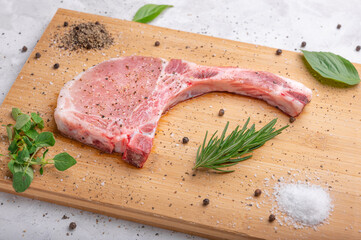 Raw Pork chop on a cutting board with herbs, rosemary, oregano, salt, pepper and basil on wooden cutting board.
