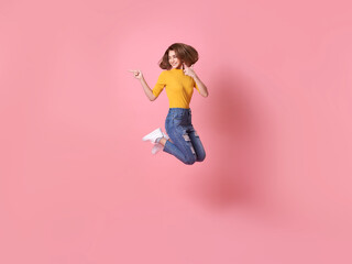 cheerful positive girl jumping in the air with raised hand pointing to copy space isolated on pink...
