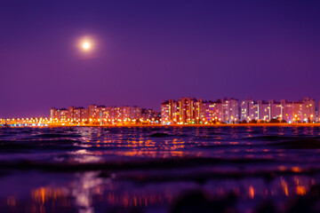 Fototapeta na wymiar Residential buildings with lanterns stand on the embankment of the river. Full moon in the night sky. Panorama of the night city. Close up view from the shore level.