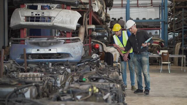 senior engineer using laptop computer checking list old used car engine with young asian worker in machinery parts shop warehouse. old manager find vehicle automobile machine in Recycle motor factory