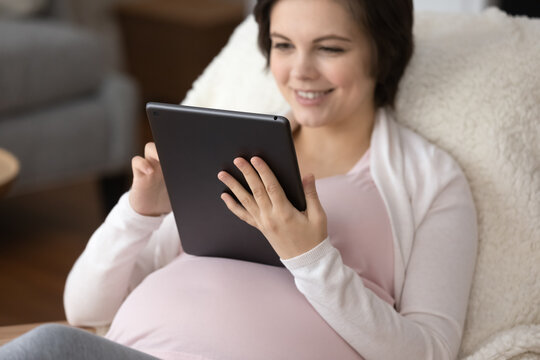 Crop close up of happy pregnant Caucasian woman relax at home talk speak on video call on pad. Smiling female rest on pregnancy maternity leave brose internet on tablet gadget. Technology concept.