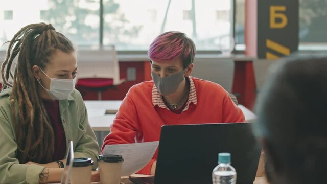 Slowmo PAN of young businesswoman with pink hair and her female colleague with dreadlocks wearing face masks and discussing work while sitting at table in office