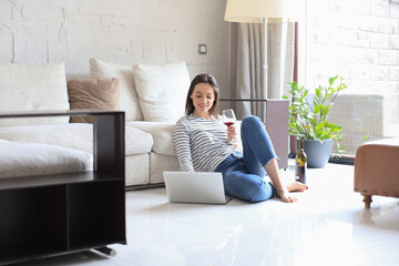 Smiling young woman sitting on floor with laptop computer and chating with friends, drinking wine.