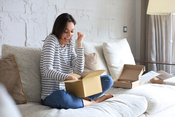 Pretty woman is unpacking parcel, expressing joy, sitting on sofa at home. - obrazy, fototapety, plakaty