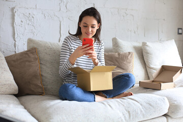 Beautiful young woman is holding cardboard box and unpacking smartphone sitting on sofa at home. - obrazy, fototapety, plakaty
