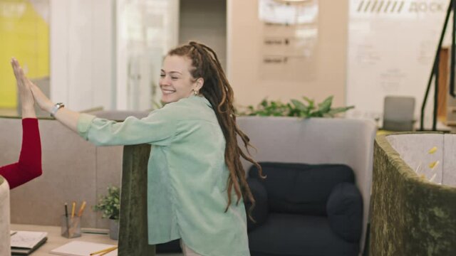 Tracking slowmo of happy young woman with dreadlocks smiling and high-fiving colleagues working in booths in start-up office