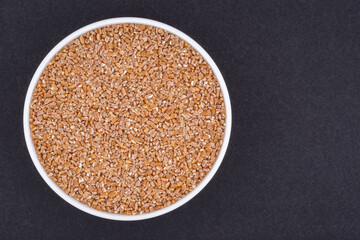 White glass bowl of wheat groats. Isolated on a dark grey background. Top view close up