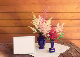 Empty white frame and red, white and pink beautiful astilbe flowers in blue vases on wooden table. Selective focus.