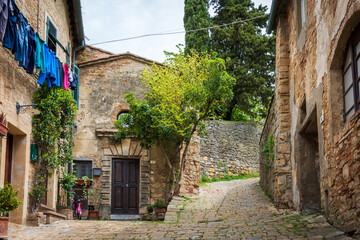 Fototapeta premium Narrow street in the old town of Volterra in a summer sunny day. Historic traditional houses and cobbled street with a tree growing on the side of an old house. Laundry is hanging from a balcony. Pisa