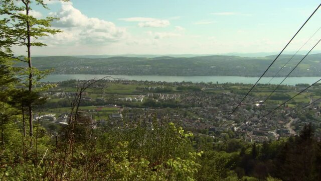 Panning Zurich And Zurichsee Lake From High On A Hill, With Rolling Hills, Bright Blue Sky, Trees, And The Cables Of The Luftseilbahn Adliswil-Felsenegg Funicular