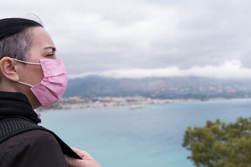 Short-haired woman dressed in black with a backpack and wearing a pink protective mask for the covid, stops on the road to look at the beautiful views towards the Mediterranean Sea.