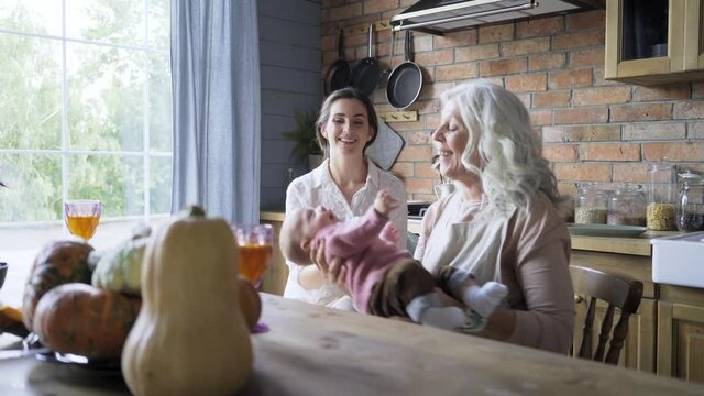 delighted grandmother plays with baby girl tossing and sitting near daughter at large wooden table with flowers wine in glasses and pumpkins at countrysside home interior