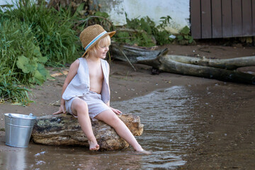 Little happy caucasian blond boy wearing straw hat sits on the tree log by the river near the metal bucket full of water. Summer kids fun theme.