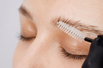 Master applies brow paste with a brush to eyebrows. Styling and lamination of eyebrows. Woman doing eyebrow permanent makeup correction. Gloved master combing patient’s eyebrows with a special brush.
