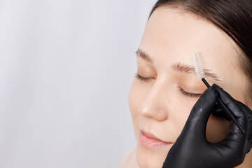 Master applies brow paste with a brush to eyebrows. Styling and lamination of eyebrows. Woman doing eyebrow permanent makeup correction. Gloved master combing patient’s eyebrows with a special brush. - obrazy, fototapety, plakaty