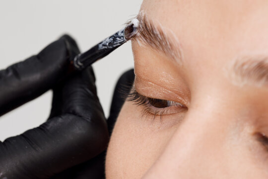 Master applies brow paste with a brush to eyebrows. Styling and lamination of eyebrows. Woman doing eyebrow permanent makeup correction. Gloved master combing patient&rsquo;s eyebrows with a special brush.