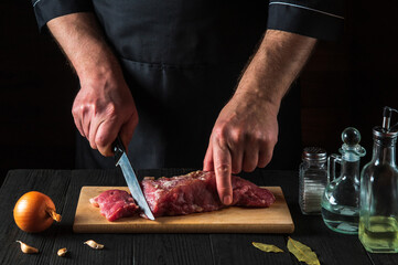 Chef or cook cutting beef meat with knife on kitchen, cooking food. Vegetables and spices on...