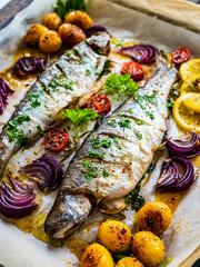 Sheet pan dinner - roasted whole trouts with lemon ,rosemary, tomatoes, red onion and potatoes on cooking pan on wooden table
