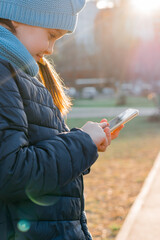 A girl in a jacket is holding a smartphone and texting a message in the rays of the evening sun. Internet surfing