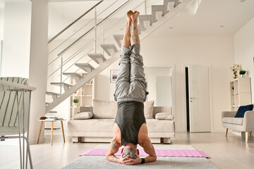 Strong fit sporty healthy old middle aged mature tattooed man doing headstand yoga morning fitness...