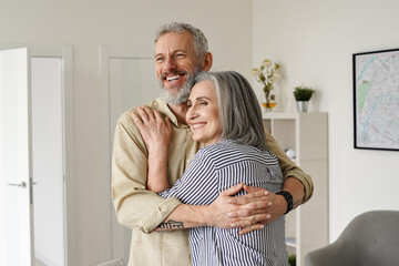 Happy senior adult mature classy couple hugging, bonding, thinking of good future. Carefree cheerful mid age old husband embracing wife looking away dreaming, enjoying wellbeing and love in new house. - obrazy, fototapety, plakaty