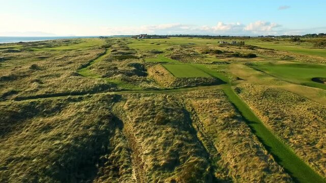 Aerial Moving Low And Forward Across The Lush Greens Of The Royal Troon Golf Club, With Blue Skies, Historic Buildings In The Distance
