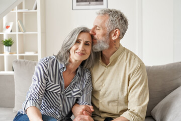 Happy affectionate classy older mature couple bonding with eyes closed at home. Loving caring senior 50s husband embracing and kissing mid aged wife enjoying tender moment sitting on couch at home.