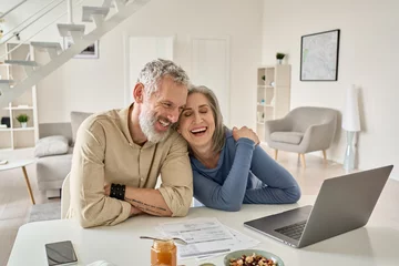 Fotobehang Gelukkig volwassen ouder familiepaar lachen, hechting aan huis tafel met laptop. Glimlachende man en vrouw van middelbare leeftijd die plezier hebben met het kopen van verzekeringen, het online betalen van rekeningen. © insta_photos