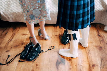 Scottish wedding preparations. Man in a kilt stands next to woman in a skirt with high-heeled shoes. There are shoes with long laces on the floor - obrazy, fototapety, plakaty