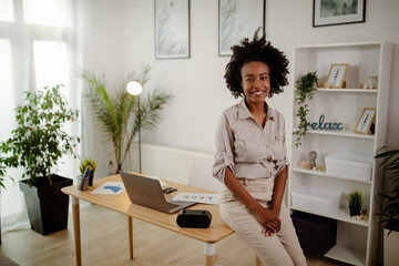 Portrait of black curly businesswoman smiling and looking at the camera while sitting on the desk.